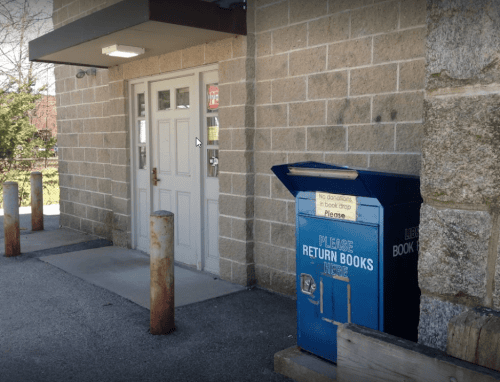 A blue outdoor library book return drop box is positioned near a beige brick building entrance with a white door and a "No donations" sign.