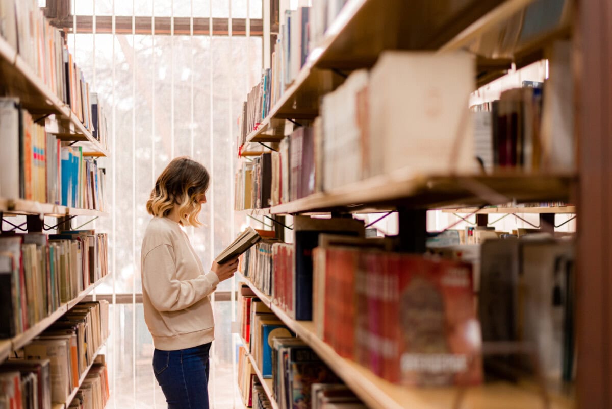 A person with shoulder-length hair stands between bookshelves in a library, reading a book.