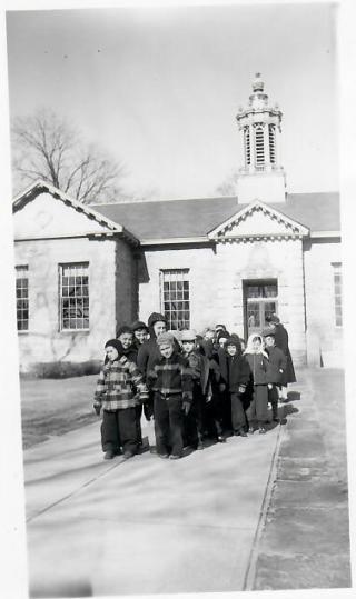 A group of children in winter clothing stand in a line outside a stone building with large windows and a cupola on the roof.