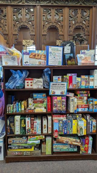 A wooden shelf filled with a variety of board games, books, and kits, with informational signs and bags displayed on top in a library or community center setting.